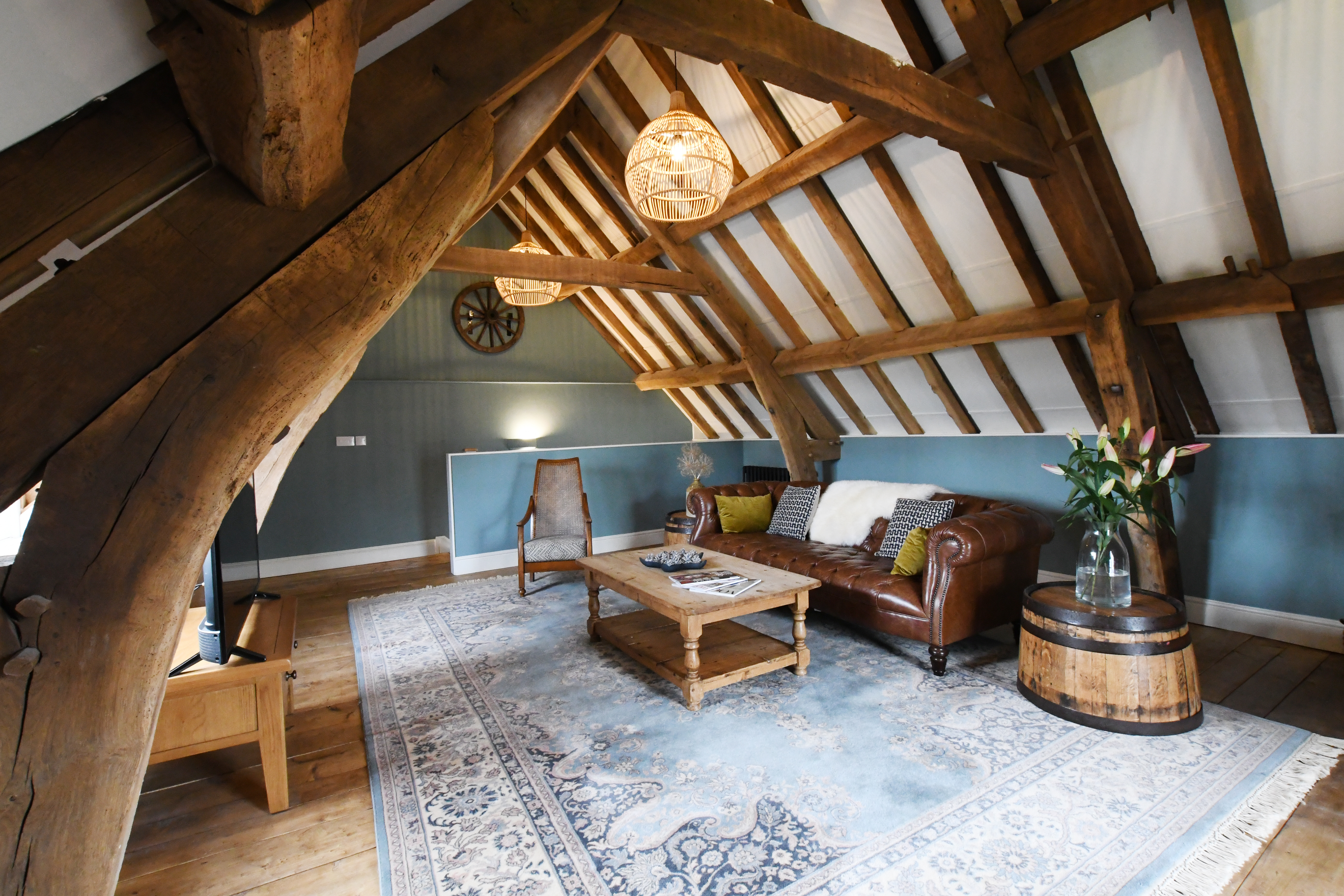 Lounge area at the top of a timber framed building, with exposed ceiling beams, a large leather sofa and wooden coffee table facing a flat-screen TV