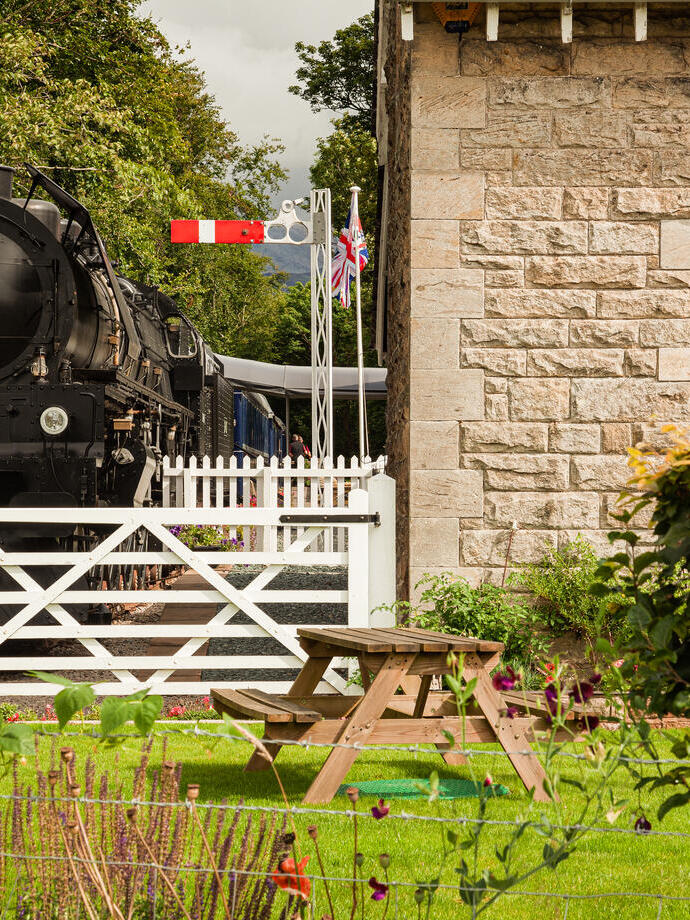 A front view of a parked steam train behind closed white level crossing gates, with a stone train station house to the right. In front is a green lawn with a picnic table. Bassenthwaite Lake Station - Silver award winner for New Tourism Business of the Year at the VisitEngland Awards for Excellence 2023.