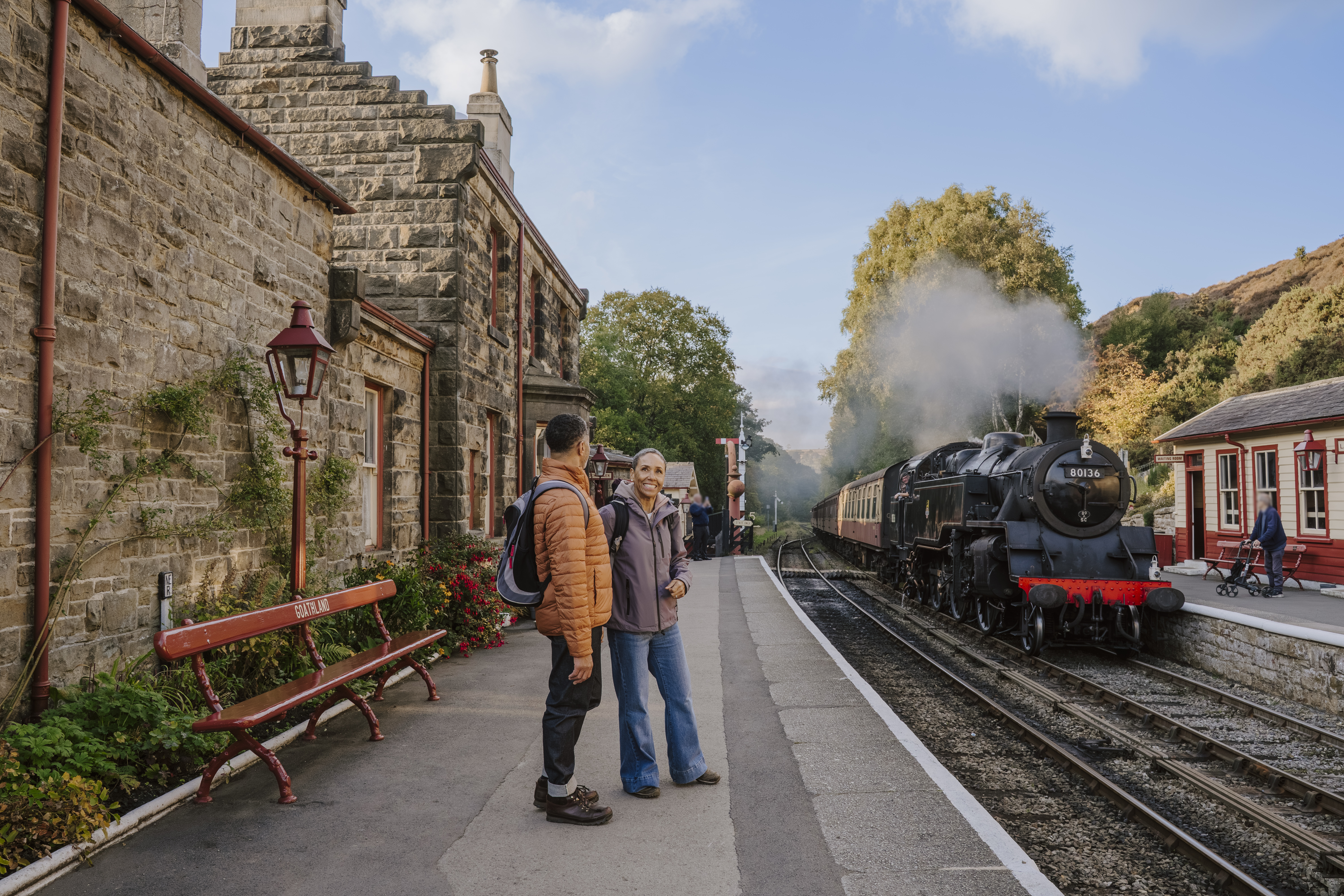 Two people stand talking on a train station platform beside an old stone building with a steam train on the tracks. Trees with autumn foliage are visible in the background.