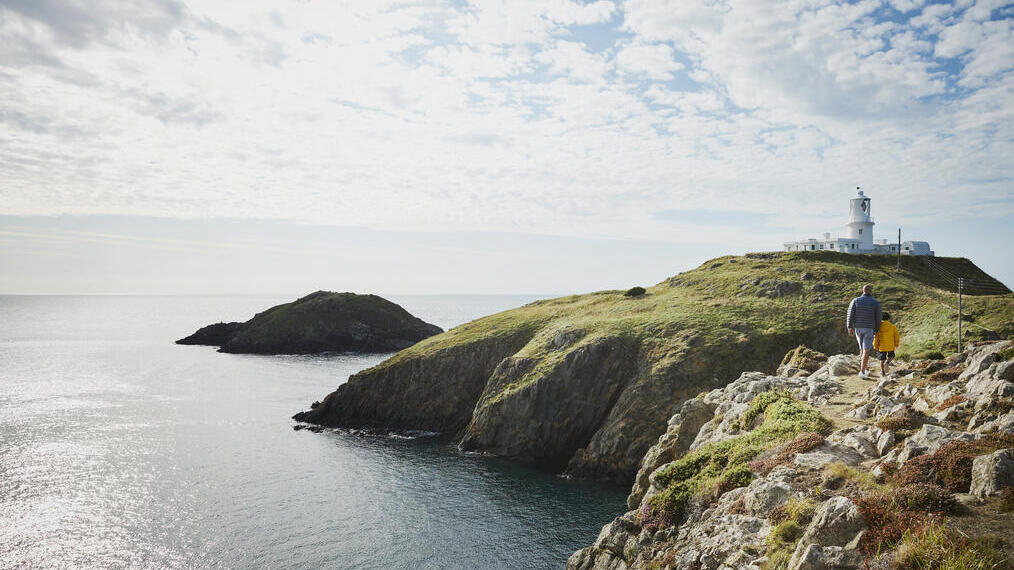 Man and child walking on a coastal path towards a lighthouse