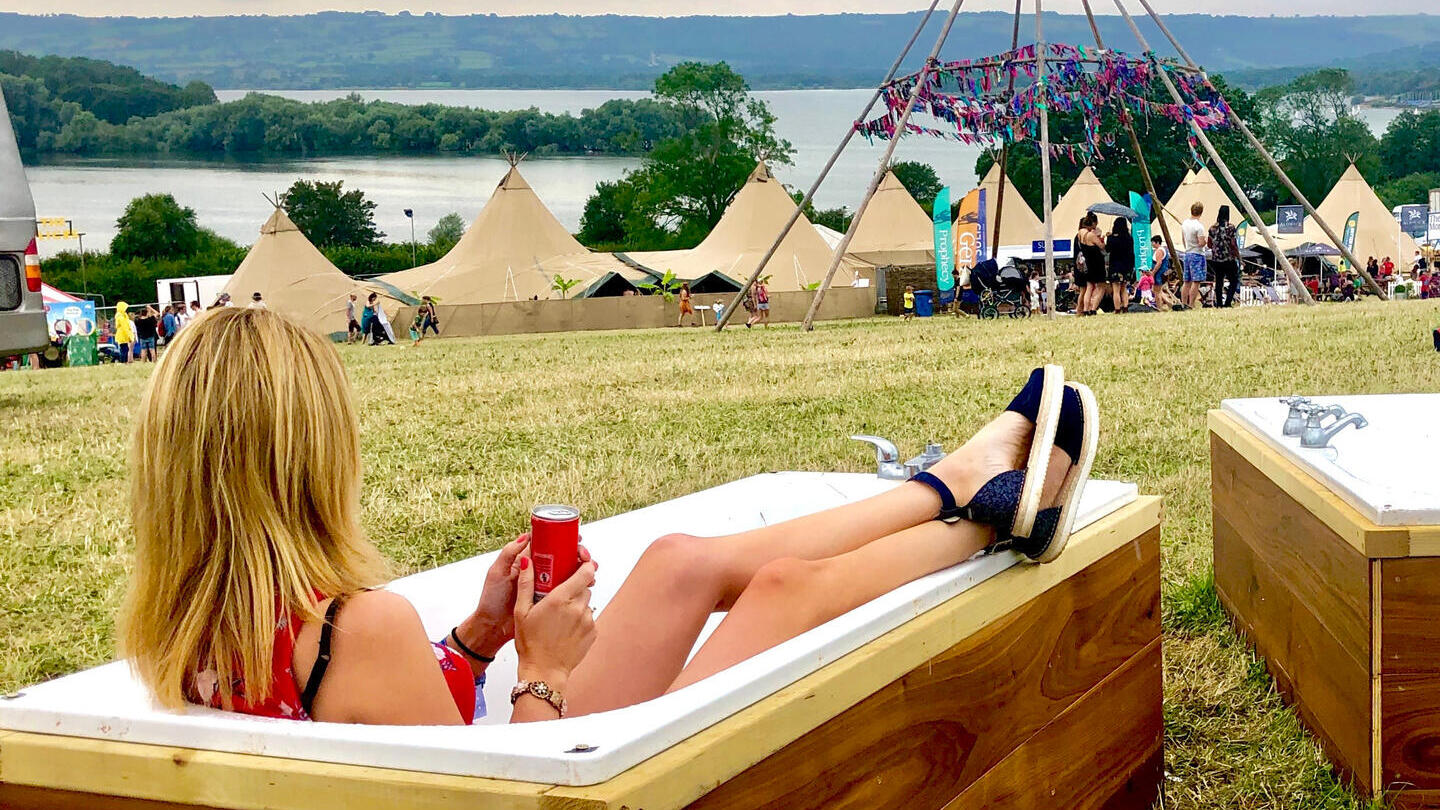 Une femme assise dans une baignoire avec vue sur les tentes du festival, les collines et un lac au loin