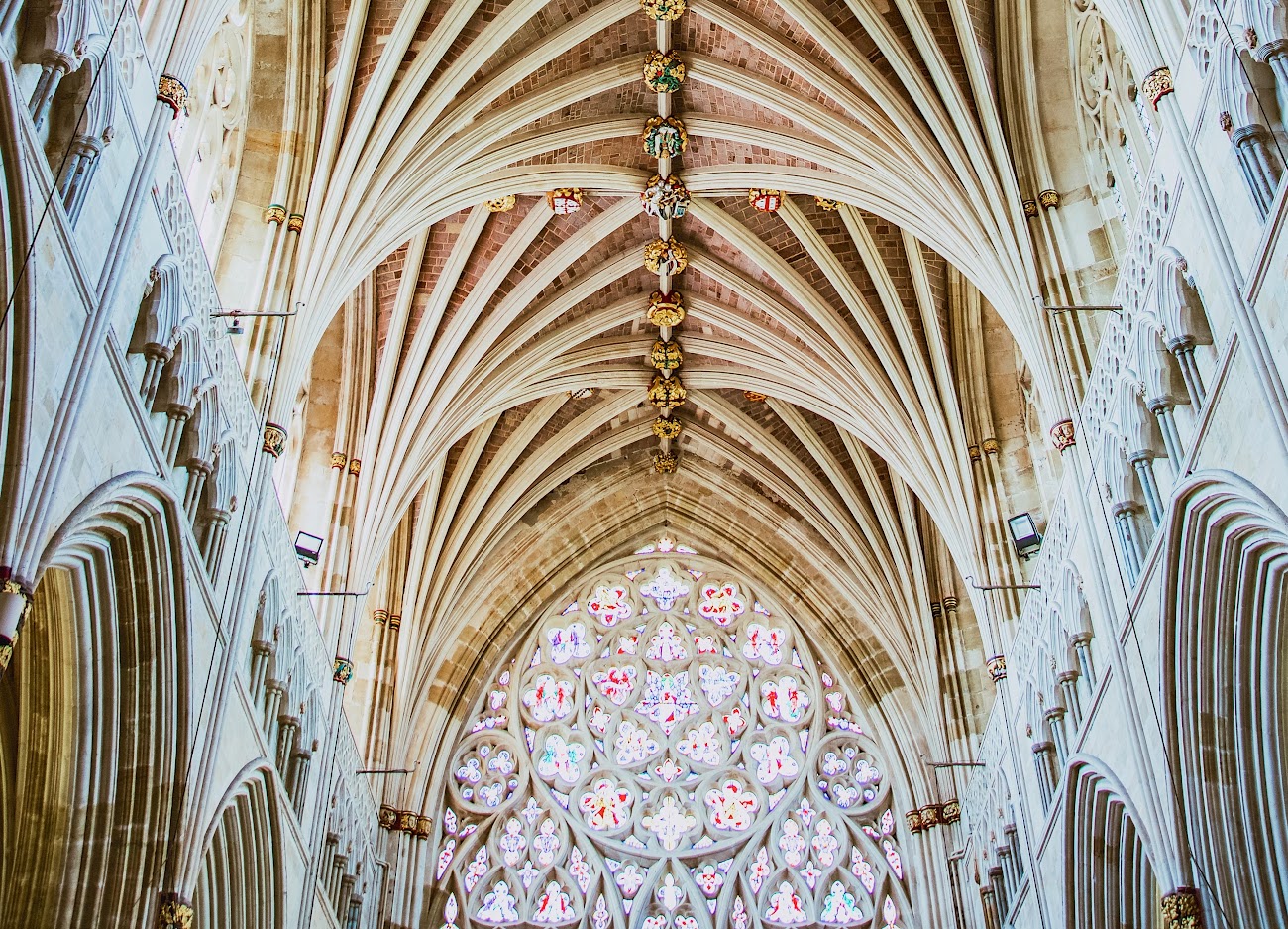 Vaulted ceiling and stained glass window of a grand Gothic cathedral interior, featuring ornate stonework and arches.