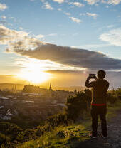 Junger Mann steht auf einer grasbewachsenen Klippe und fotografiert den Sonnenuntergang