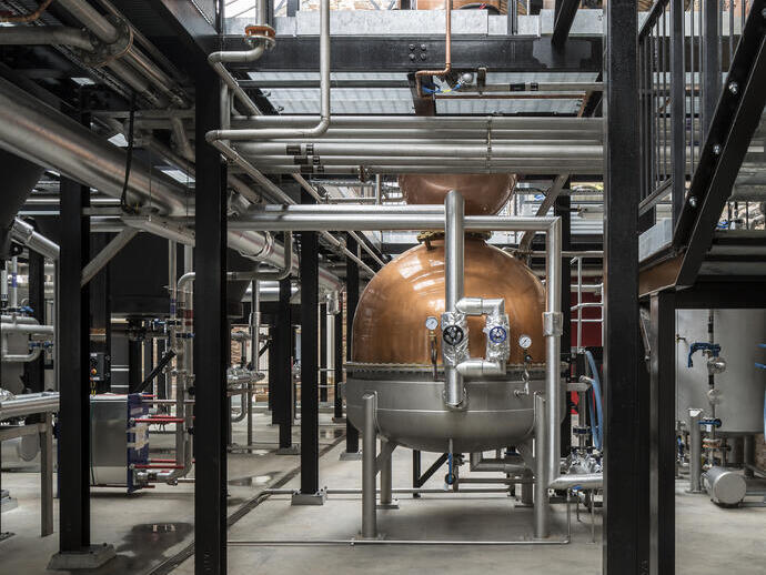 Industrial distillery with metal stairs, copper stills, pipes, and tanks inside a high-ceilinged building.