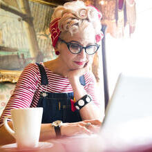 Close up of a blond woman wearing a stripy top and red head scarf, having a coffee and looking down at a lap top screen, smiling.