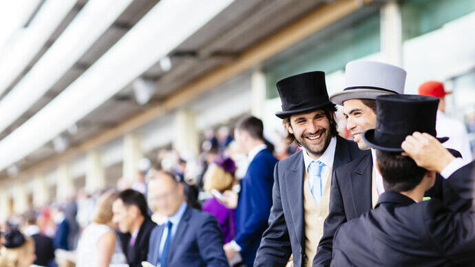 Twee mannen die glimlachen in formele kleding op een drukke tribune bij een paardenrenbaan