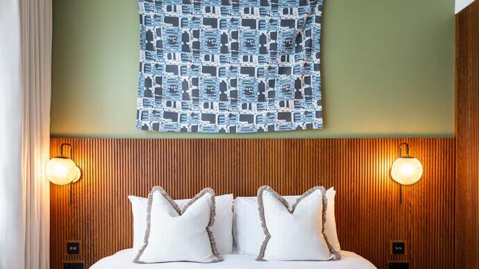 Modern hotel room with a double bed, two white pillows, wooden headboard, wall art, and two round wall lamps on either side of the bed.