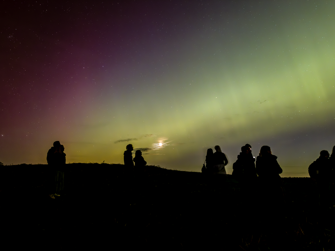 Silhouetted group of people watching the northern lights in a dark landscape under a colorful night sky.
