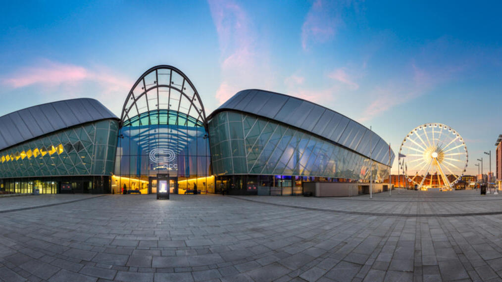Modern glass building with curved roof at sunset, open plaza, and a large Ferris wheel in the background, surrounded by cityscape elements.