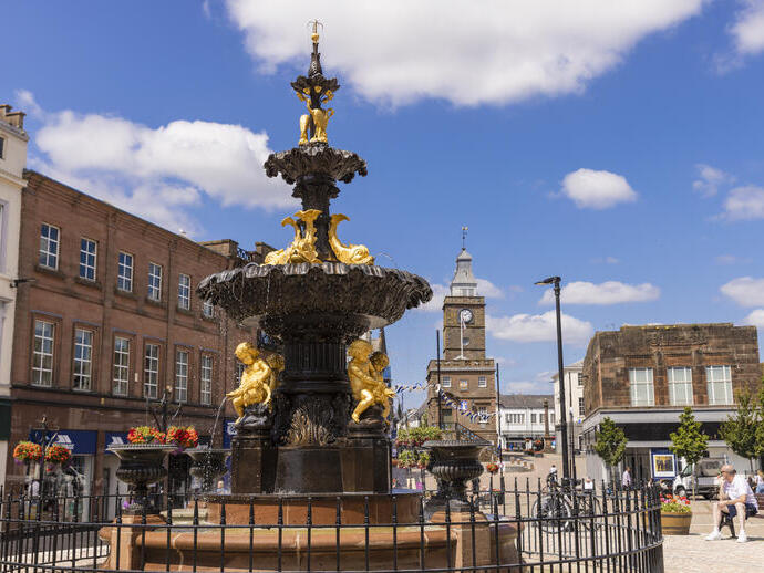 Una ornamentada fuente negra y dorada con estatuas de personas y peces en el centro de una calle comercial.
