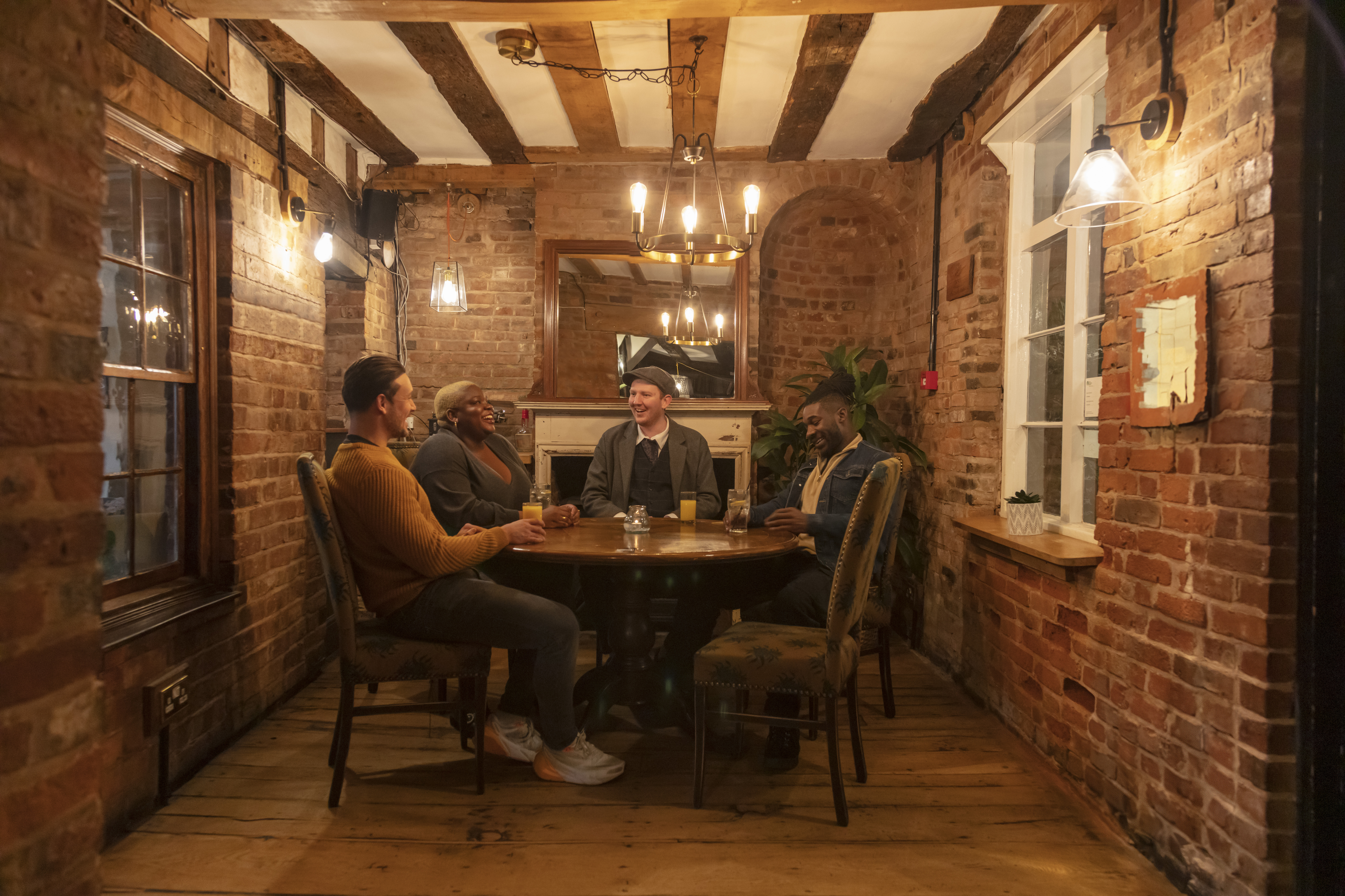 Four people sit around a round table in a cozy, rustic brick-walled room with wooden beams, warm lighting, and a relaxed atmosphere.
