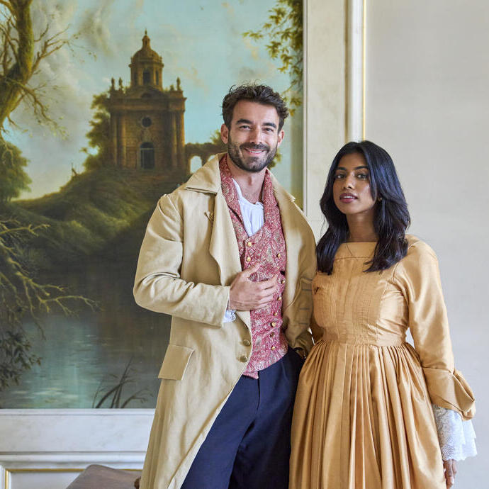 Ein Mann und eine Frau in historischen Kostümen stehen in einem dekorierten Raum vor einem Gemälde mit Landschaft und altem Gebäude.