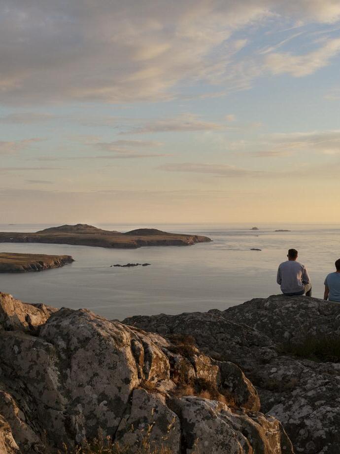 Zwei Männer sitzen auf einer Felsklippe und blicken in der Abenddämmerung auf das Meer
