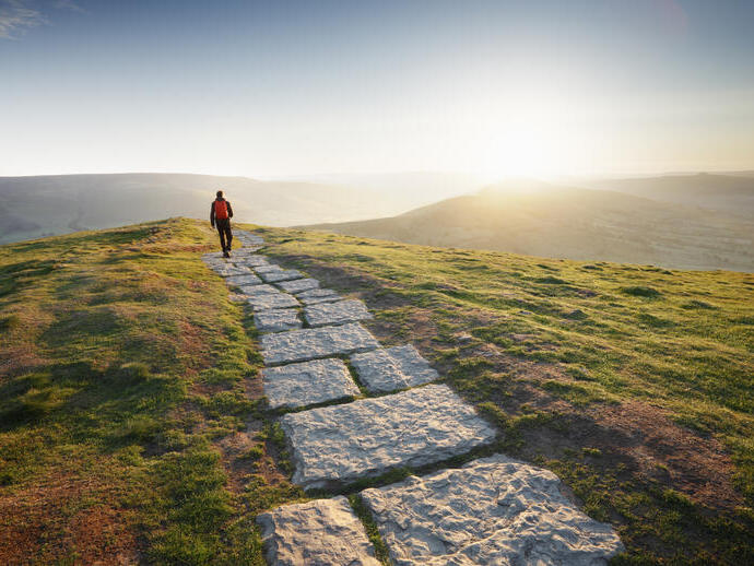 Man in red jacket walking away into the sun at the top of a mountain path