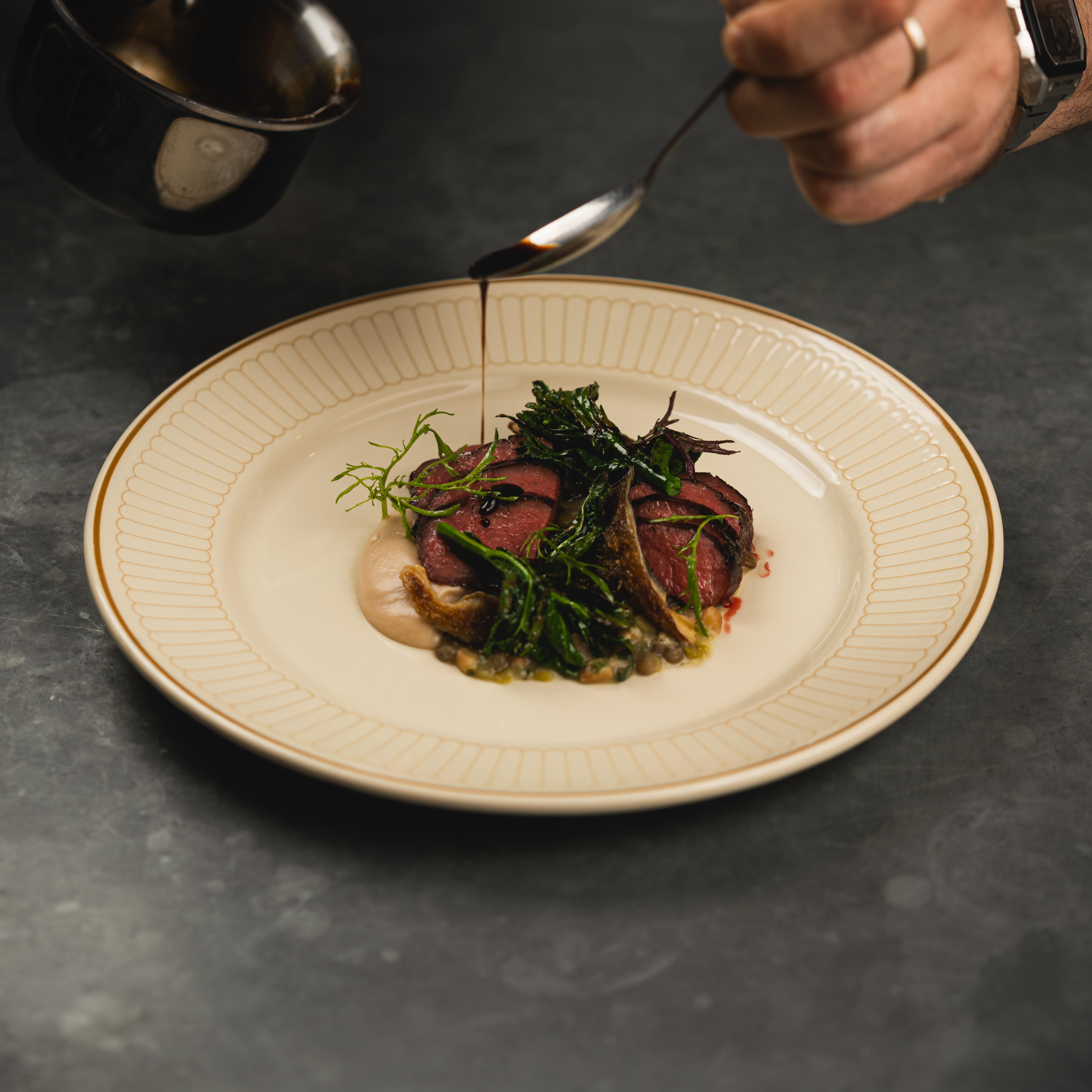 Plated gourmet dish being garnished with sauce, featuring sliced meat, greens, and a creamy puree on a patterned plate.