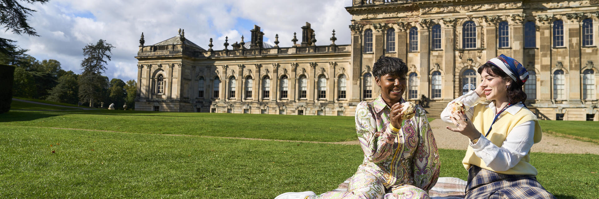 Zwei Personen sitzen auf einer Decke auf dem Rasen vor einem großen historischen Gebäude.