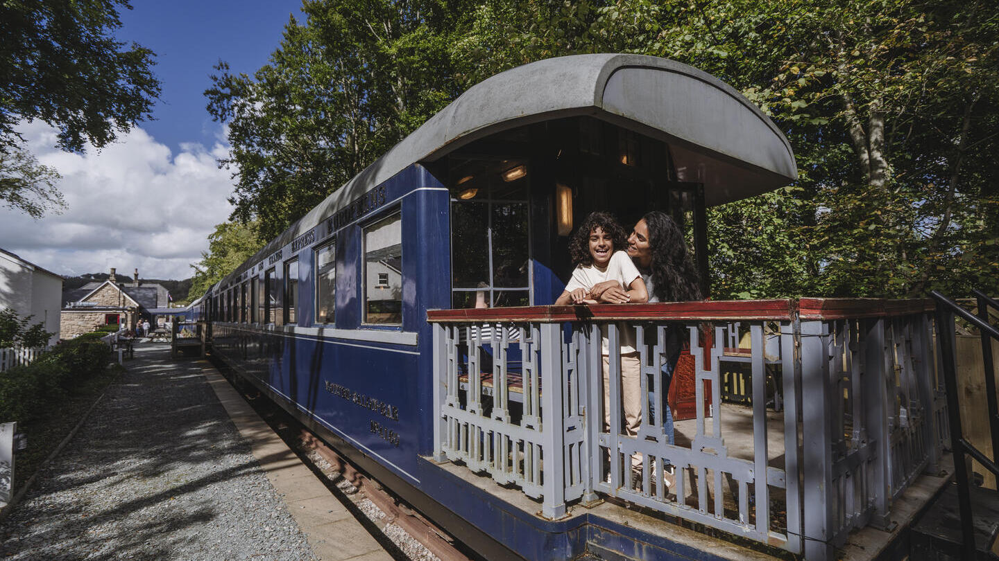 Historic blue train carriage at a station with trees around, people standing on the train's outdoor platform, and a house in the background on a sunny day.