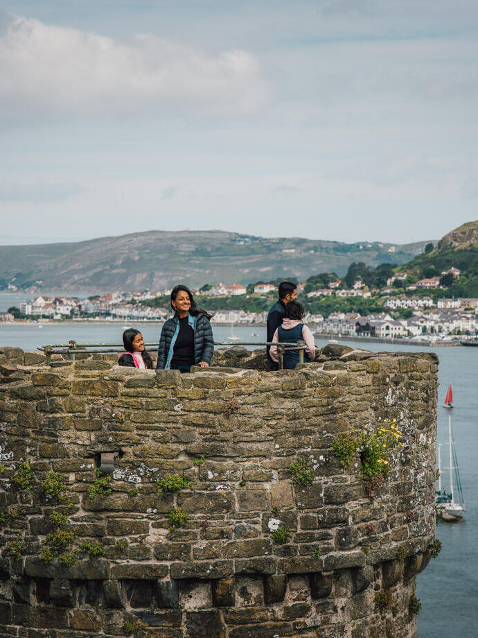 People standing atop an old stone castle tower overlooking water, sailboats, and a coastal town with hills in the background under a cloudy sky.