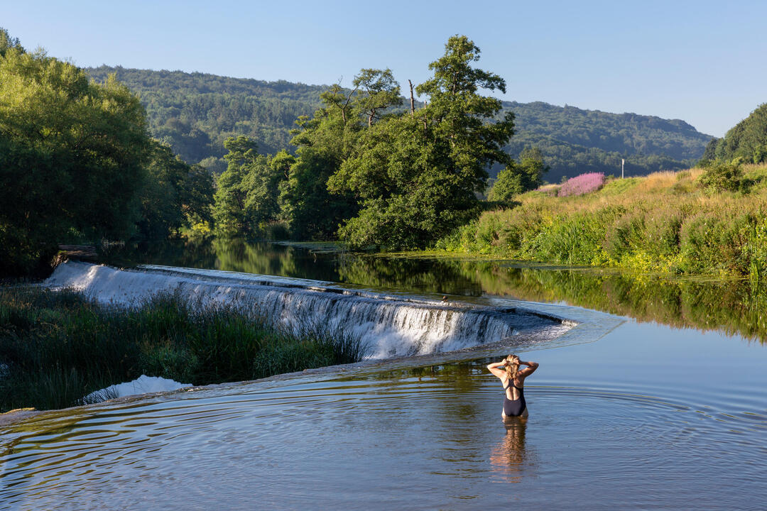 A female swimmer standing in the shallows preparing to swim in the a river above a weir with wooded hills in the distance 