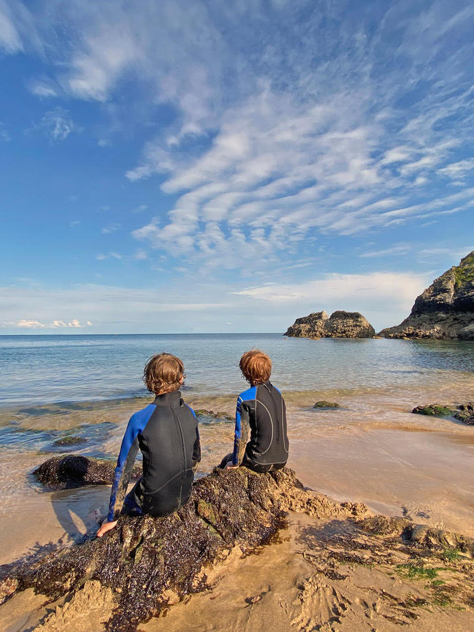 Back view of two young boys in wetsuits sitting on a rock looking out to sea with blue sky