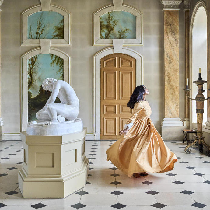 Mujer con vestido largo girando en un gran salón con escultura de mármol, columnas y paredes decoradas.