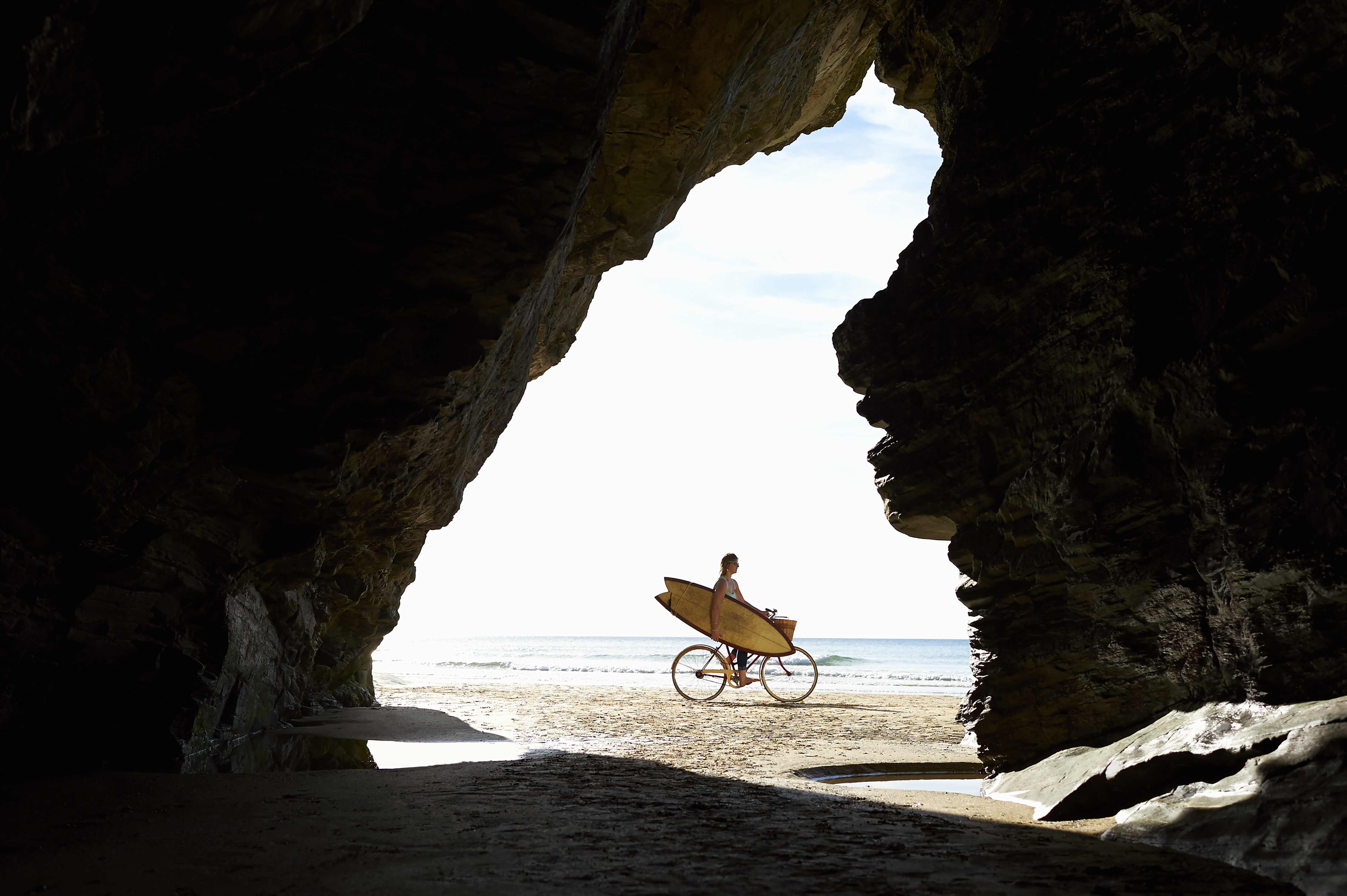 Persona en bicicleta con tabla de surf en la playa, vista desde el interior de una cueva.