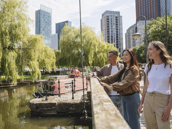 Three people stand on a canal bridge with modern high-rise buildings, green trees, and a pink canal boat in the background.