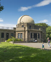 Observatory with a domed roof surrounded by greenery under a blue sky, with people walking in the foreground.