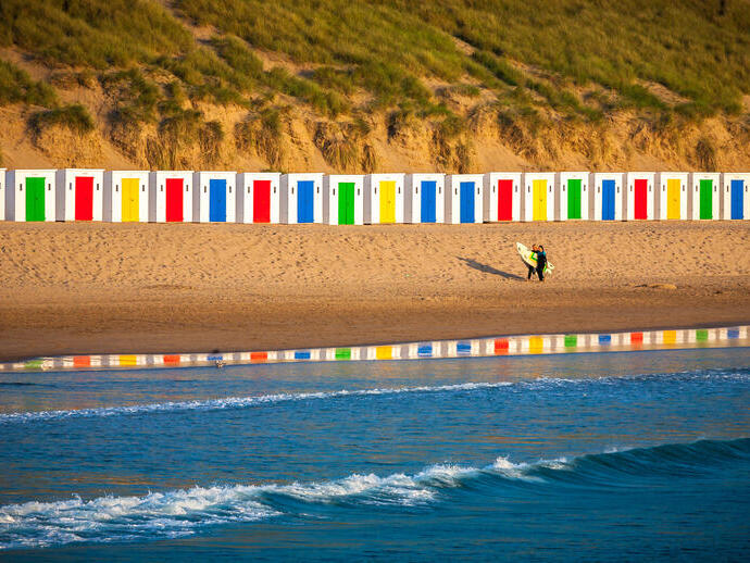 Un surfista caminando por la playa frente a coloridas casetas de playa.