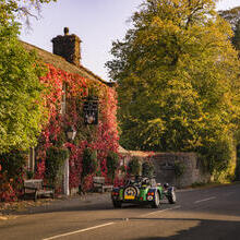 A classic car drives past a pub covered with ivy leaves