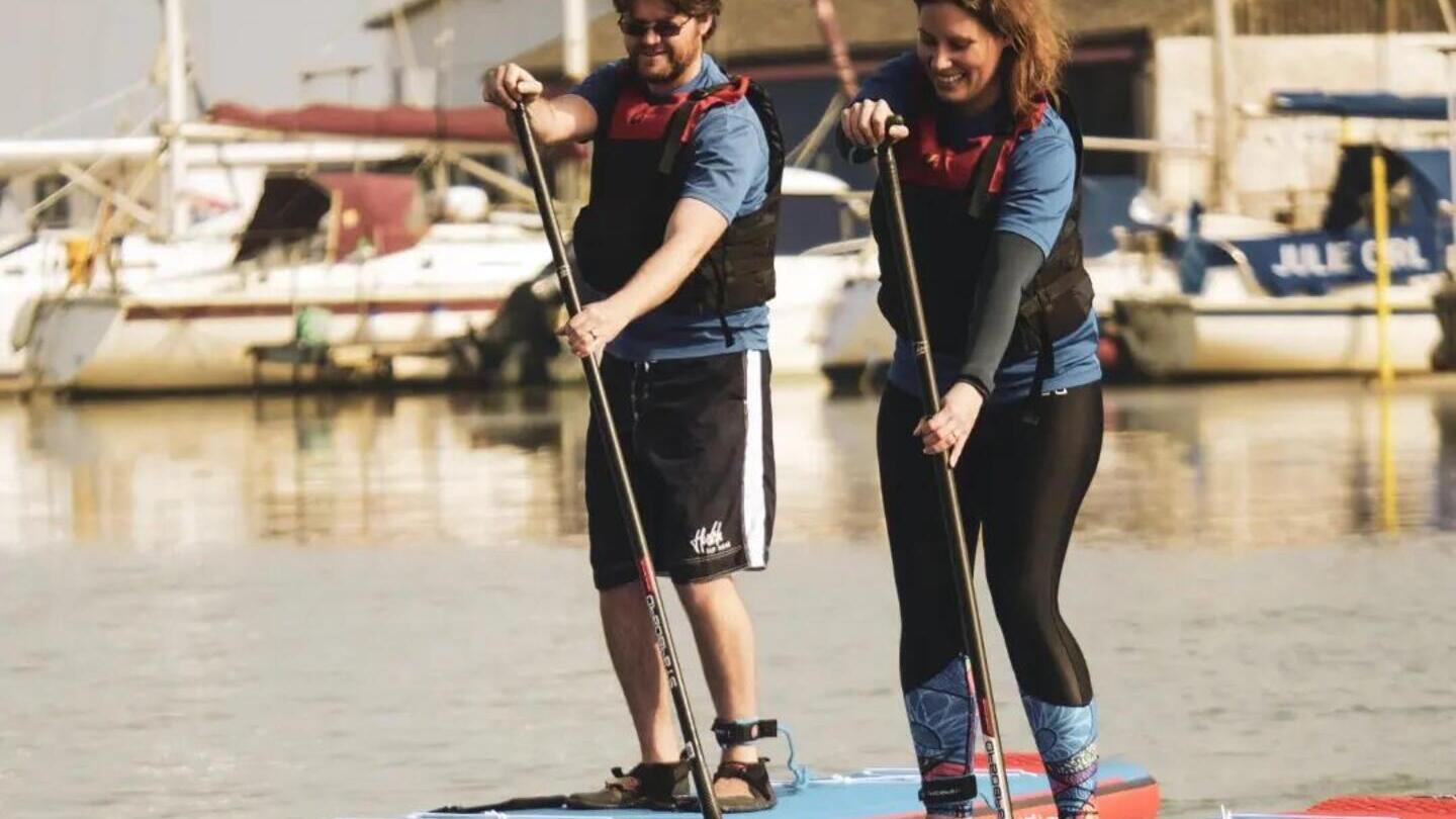 Hombre y mujer en tablas de paddle surf con barcos al fondo