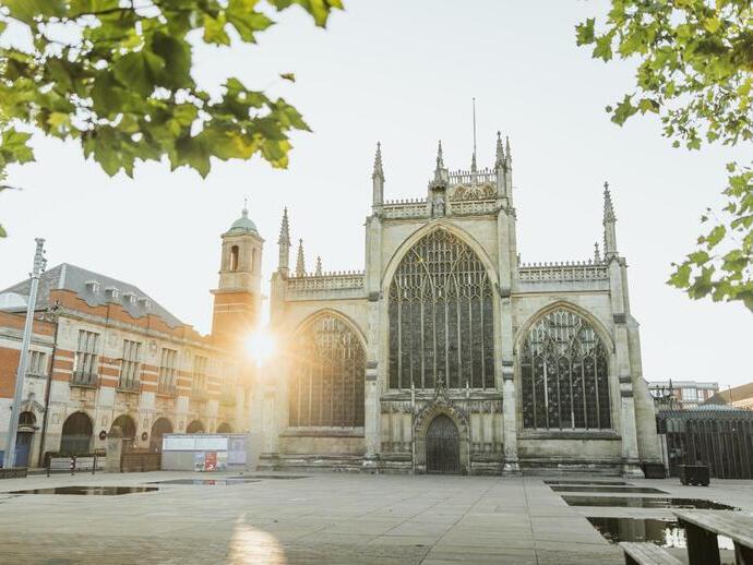 Historic church with large stained glass windows on a city square, sunrise in the background and leafy trees framing the scene.