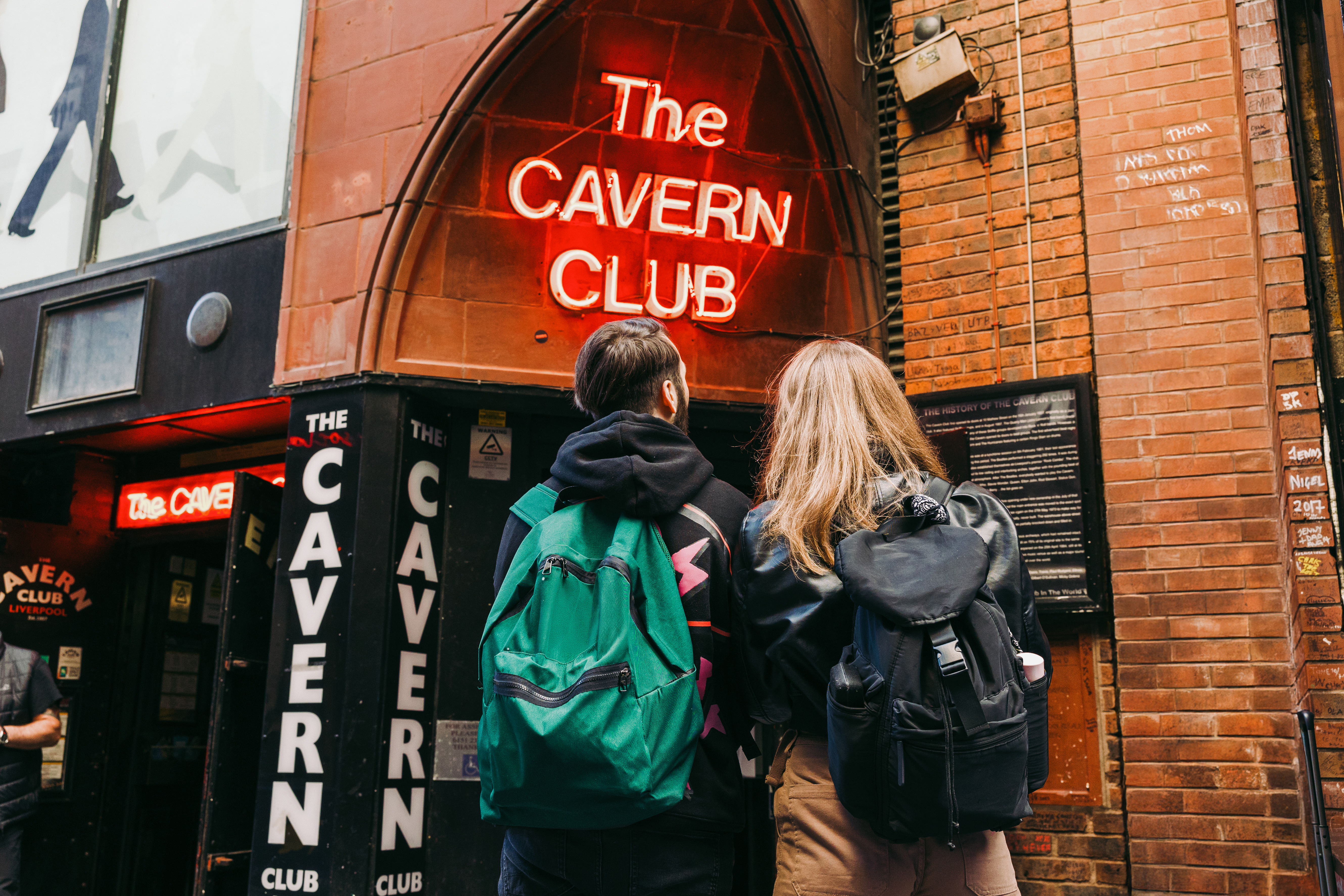 Two people with backpacks stand outside The Cavern Club, a famous brick music venue with neon signage in Liverpool.
