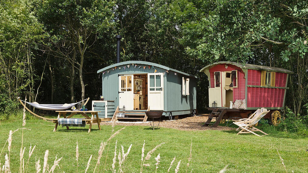 Colourful shepherd's hut and gypsy caravan style accommodation in a tree-edge field with a hammock and picnic table in front