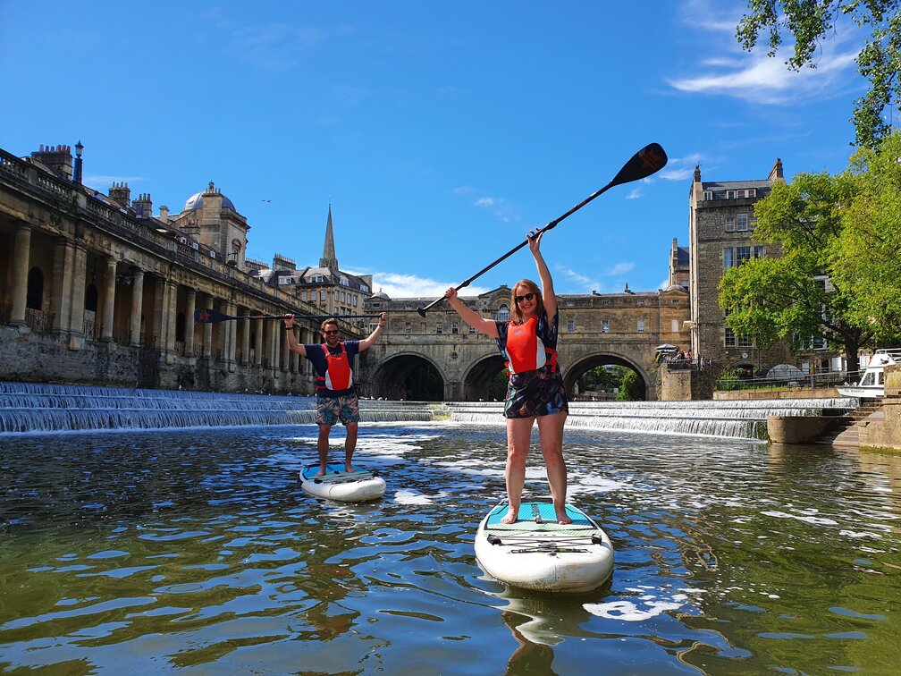 Two people paddleboarding down a river in the centre of Bath
