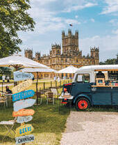 Un food truck, ombrelloni e cartelli indicatori nel parco di un castello