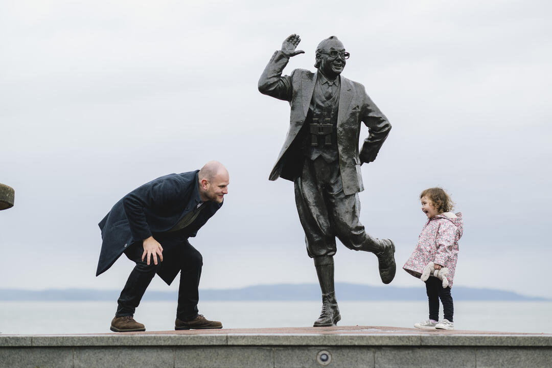 A man and young child interact playfully with a statue on the seafront
