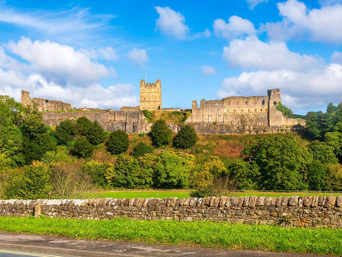 Historic stone castle ruins on a hill with green trees and a stone wall under a blue sky with scattered clouds.