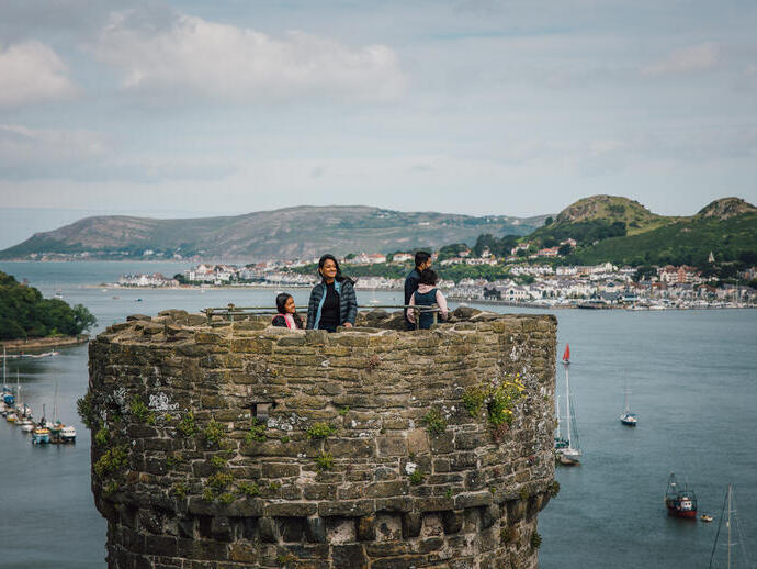 People standing atop an old stone castle tower overlooking water, sailboats, and a coastal town with hills in the background under a cloudy sky.