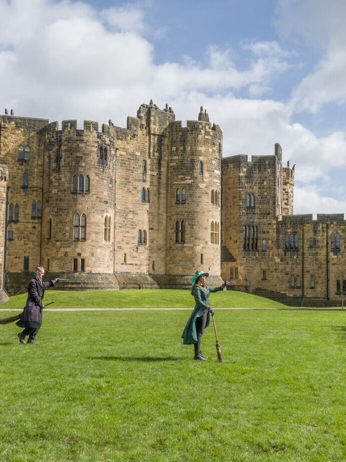 A group of people lined up for a Broomstick Training lesson with the wizarding professors by a large castle.
