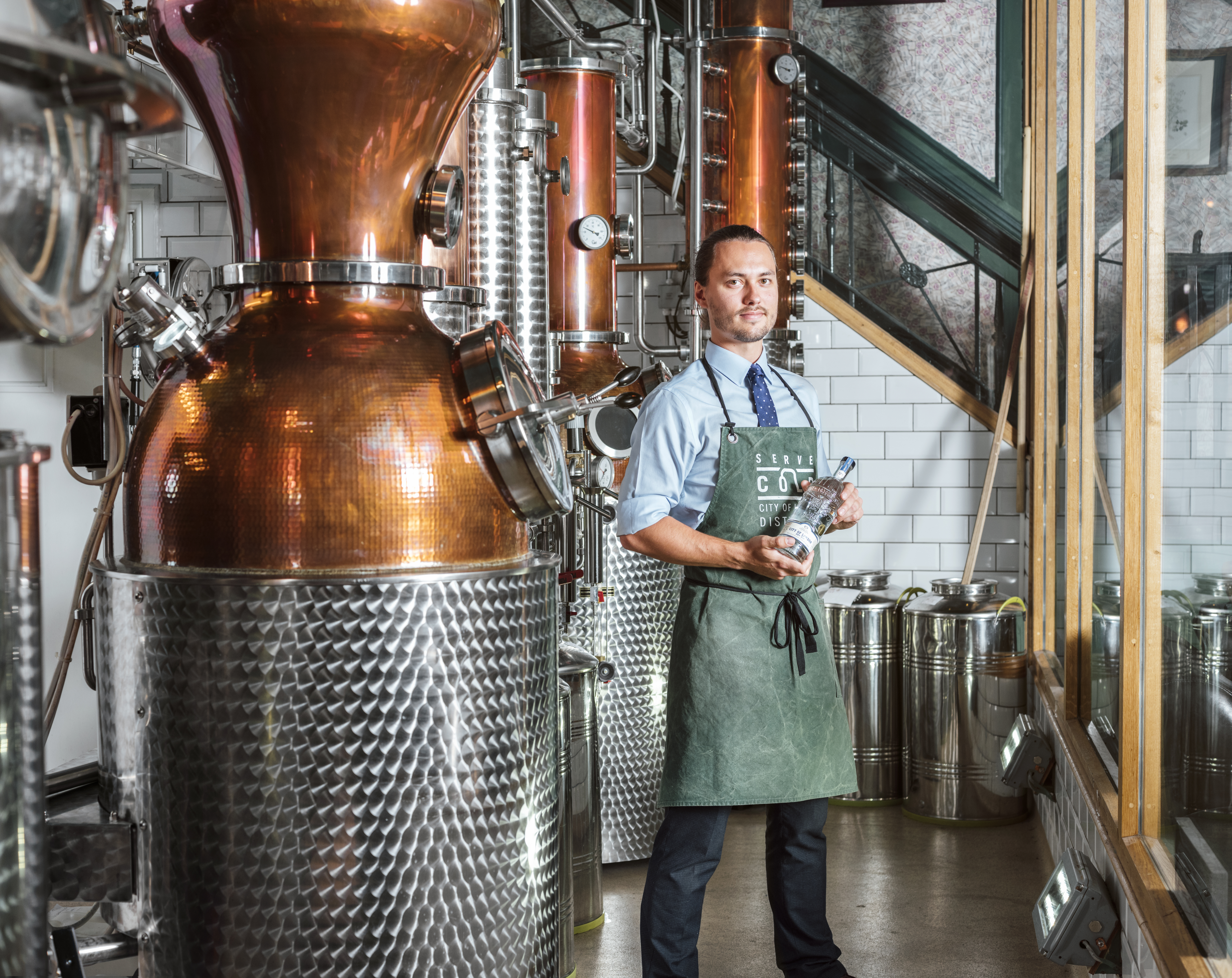 Man wearing green apron standing next to a copper still in a gin distillery