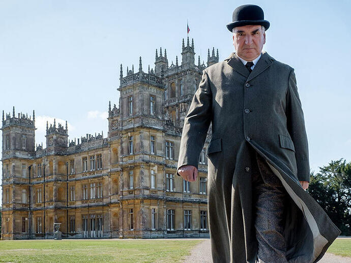 Une personne en tenue d'époque se tient devant un grand bâtiment historique à tours, entouré de jardins paysagers par temps clair.
