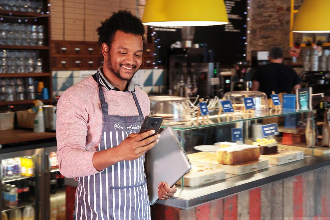Male cafe owner smiling at his phone in front of the counter in the restaurant