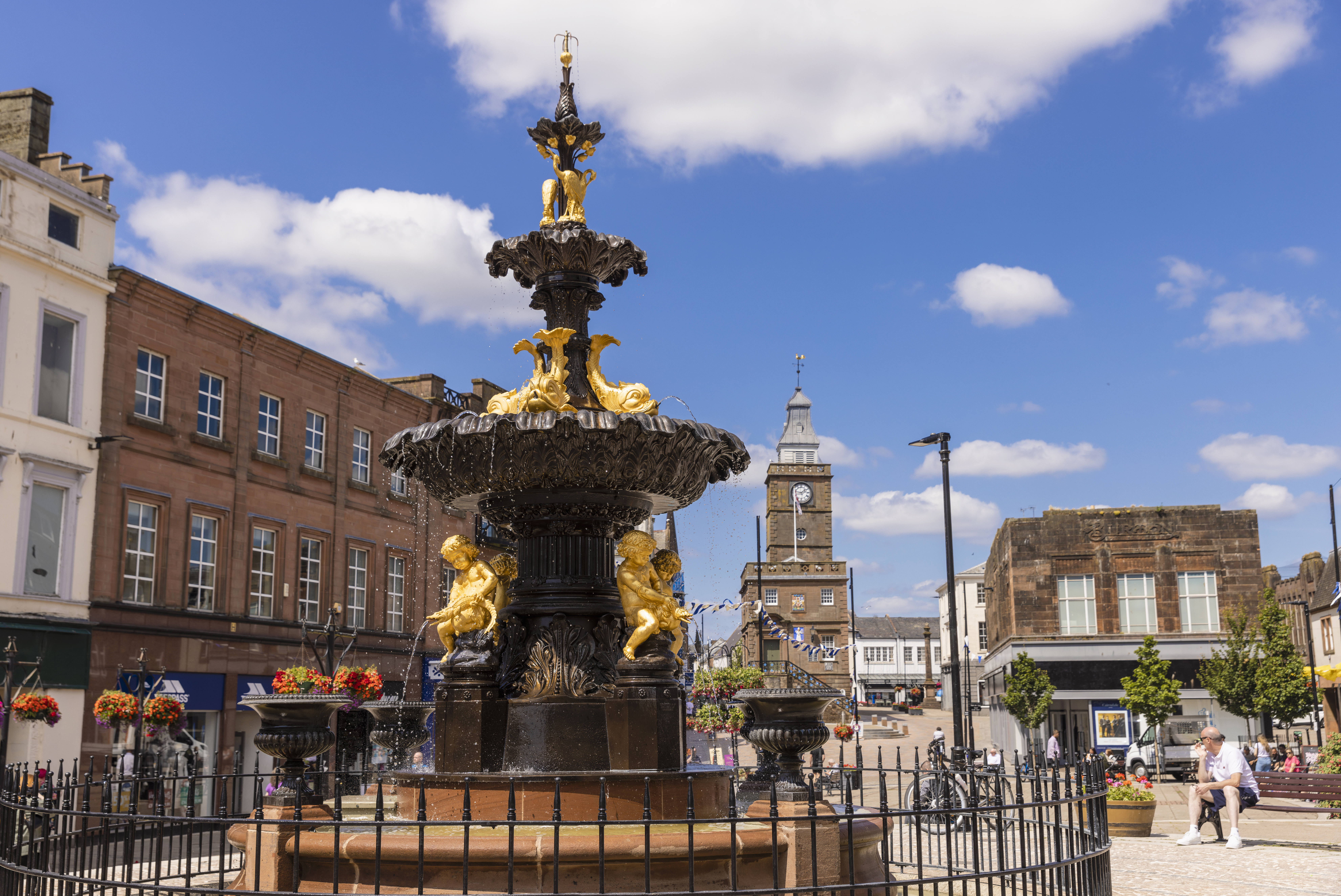 An ornate black and gold fountain with statues of people and fish in the centre of a shopping street.