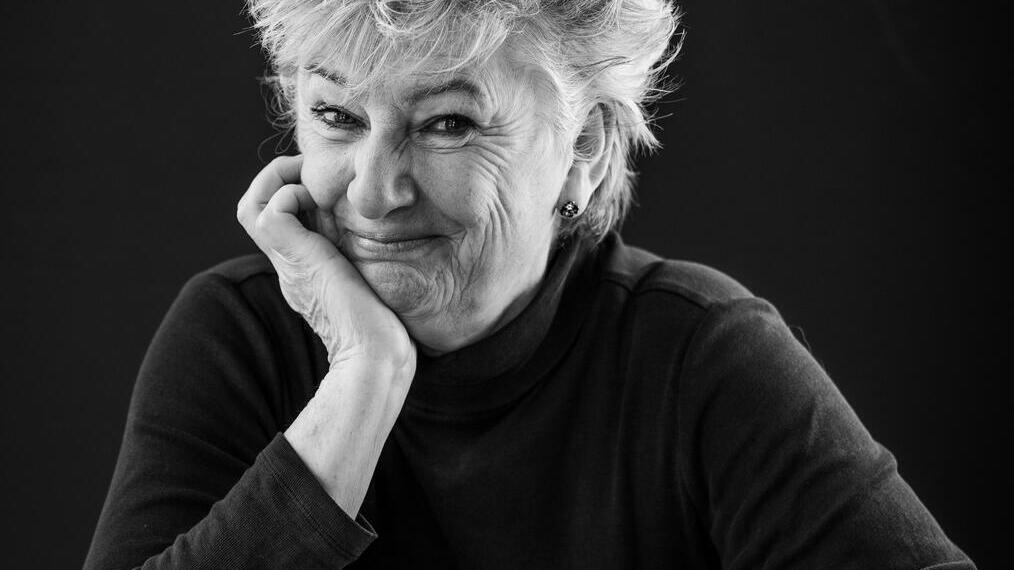 Black and white portrait of an older person smiling, seated at a table, with a dark background.