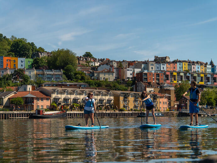 Gente practicando paddle surf en el río con la ciudad de Bristol al fondo.