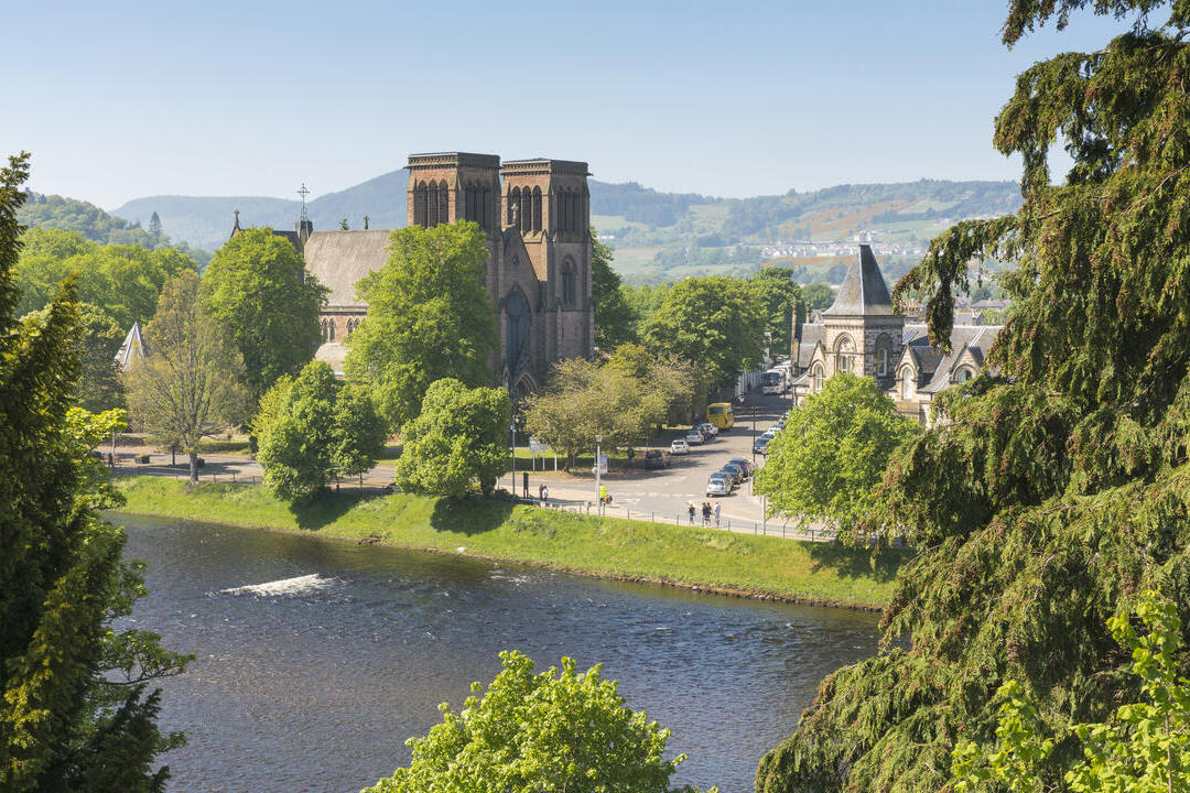 A cathedral with two towers overlooks a river on a sunny day, with trees in the foreground.