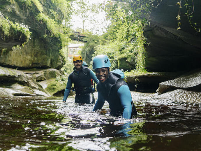 Two men wearing wetsuits and helmets wade waist deep in a gorge