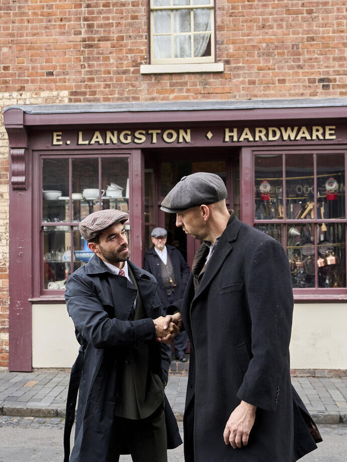 Two men in vintage attire shake hands outside a brick hardware store named E. Langston. Historical street scene with older shopfronts.