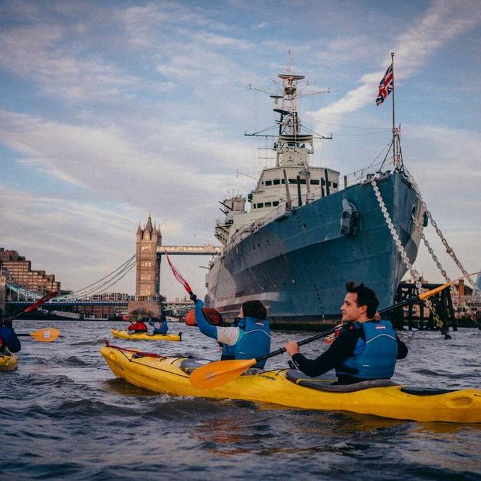 People in Kayaks on a river past iconic navy ships and iconic city landmarks.